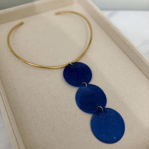 Può includere: Una collana girocollo dorata con tre pendenti circolari blu reale. La collana è esposta su una superficie color crema all'interno di un vassoio bianco. I pendenti sono collegati con piccoli anelli d'argento.