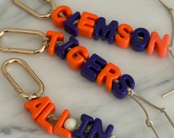 May include: Three keychains featuring gold-toned metal lobster clasps attached to beige cords. The cords are strung with orange and purple plastic letter beads spelling out CLEMSON, TIGERS, and ALL IN, with a single white bead between ALL and IN.