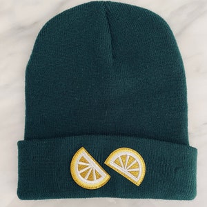 Może przedstawiać: Czapka w ciemnozielonym kolorze z dwoma złotymi naszywkami w kształcie plasterków cytryny z przodu.