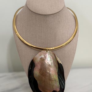 Könnte beinhalten: Eine goldfarbene Halskette mit einem großen, schillernden Anhänger. Der Anhänger ist eine ovale Muschel mit schwarzen, rosa und perlmuttartigen Farbtönen. Die Halskette wird auf einer neutralfarbenen Schaufensterpuppe präsentiert.