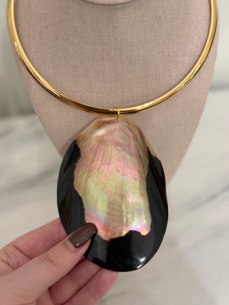 May include: A gold-tone choker necklace with a large, iridescent pendant. The pendant is a mix of black and pearlescent colors, with a shell-like appearance. The necklace is displayed on a neutral-colored mannequin.