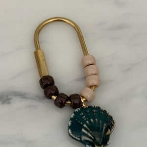 May include: A gold-toned keychain with a dark teal seashell pendant. The keychain features a U-shaped design with a cylindrical gold-toned base and a series of brown and pink beads. The seashell pendant is detailed with white accents.