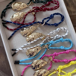 May include: A selection of rope bracelets in various colours, each with a gold-coloured, textured charm. The bracelets feature combinations such as green, red, blue, white, light blue, pink, and yellow. Each has a knotted design and black tips.