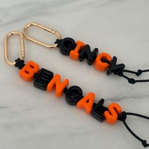 Puede incluir: Dos llaveros con mosquetones de metal dorado unidos a cordones negros. Los cordones están ensartados con cuentas de letras de plástico naranja y negro que forman las palabras BENGALS en uno y CINCY en el otro, creando un accesorio deportivo.