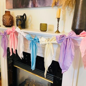 May include: A garland of five satin bows in pink, white, light blue, cream, and lavender hangs over a fireplace. The bows are tied in a simple knot and are evenly spaced.
