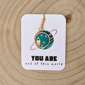 May include: A gold charm featuring a rocket ship and stars on a blue background. The charm is attached to a gold chain and hangs from a card that reads "You are out of this world".