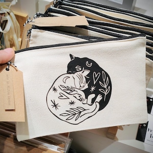 May include: Cream-colored canvas zippered pouch featuring a black and white cat design. The design shows two cats, one black and one white, intertwined with celestial and botanical elements. The pouch has a black zipper and a small tag.