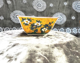 May include: A yellow, trapezoid-shaped ceramic bowl with a floral design. The bowl features white and blue flowers with green leaves against a textured yellow background. The rim is red, and the base is green. The bowl is likely for decorative or serving purposes.