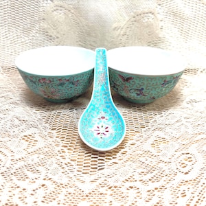 May include: A turquoise ceramic spoon and two matching bowls. The spoon and bowls have a floral design with pink, red, and yellow accents. The bowls and spoon are on a white lace tablecloth.