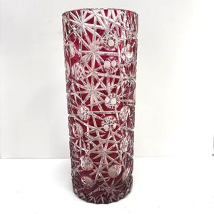May include: A tall, cylindrical vase with a ruby red interior and a clear, intricate cut glass design. The pattern features geometric shapes, including stars and circles, creating a textured surface. The vase is likely for decorative purposes.