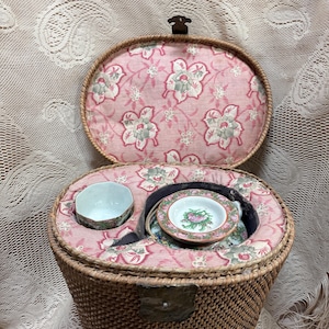 May include: A vintage wicker picnic basket with a hinged lid, lined with pink floral fabric. Inside, it holds a small teacup, saucer, and other dishware. The basket is brown with a metal clasp.