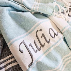 May include: A light blue and white striped towel with the name "Julia" embroidered in grey script. The towel has a fringe edge and is folded, showing the soft texture of the fabric. The towel is a light blue colour with white stripes.