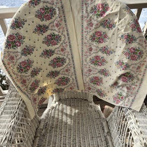 May include: A white wicker chair with a floral patterned tablecloth draped over it. The tablecloth has a cream background with pink, blue, and green floral designs.