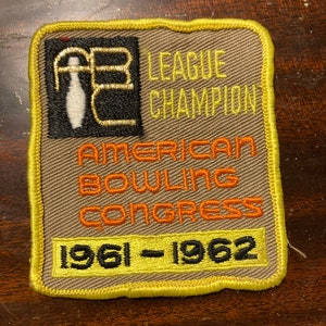May include: A tan and yellow embroidered patch with the text "League Champion American Bowling Congress 1961-1962". The patch features a black and gold bowling pin and ball graphic.