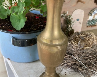 May include: A gold brass vase with a flared top and a round base. The vase has a smooth, polished finish and is sitting on a white wooden surface.