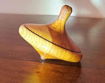 May include: A wooden spinning top with a light brown and dark brown wood grain pattern. The top is resting on a dark brown wooden surface.