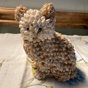 May include: A small, white and brown cat figurine made entirely of seashells. The cat has black eyes and is sitting on a white tablecloth with floral designs.
