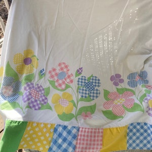 May include: A white bed sheet with a floral pattern in shades of pink, yellow, blue, and purple. The flowers are arranged in a row along the bottom edge of the sheet. The sheet has a patchwork border with squares of different colors and patterns.