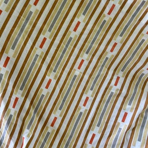 May include: A white fabric with a repeating pattern of vertical stripes in shades of brown, gray, and yellow. Small orange rectangles are interspersed along the stripes. The fabric is slightly wrinkled, suggesting it is a sheet or other textile.