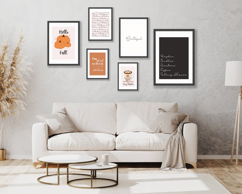May include: A collection of framed art prints with fall-themed designs. The prints feature text like "Hello Fall" and "Grateful", along with images of pumpkins and fall-related items. The frames are black, and the art is displayed above a white sofa.