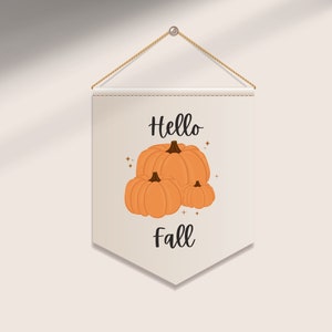 May include: A beige pennant banner with the words "Hello Fall" and an illustration of three orange pumpkins. The banner is suspended by a natural rope and a metal eyelet, perfect for autumn decor.