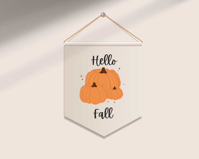 May include: A beige pennant banner with the words "Hello Fall" and an illustration of three orange pumpkins. The banner is suspended by a natural rope and a metal eyelet, perfect for autumn decor.