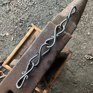 May include: A silver metal chain with decorative loops, resting on a brown metal anvil.