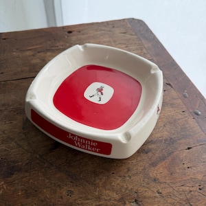 May include: Cream-colored ashtray with a red center featuring a logo of a man in a red coat. The ashtray has a rectangular shape with rounded corners and the words "Johnnie Walker" in white.