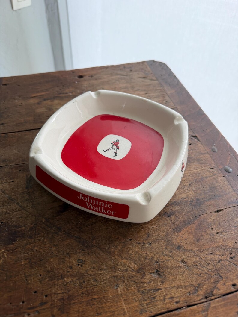 May include: Cream-colored ashtray with a red center featuring a logo of a man in a red coat. The ashtray has a rectangular shape with rounded corners and the words "Johnnie Walker" in white.