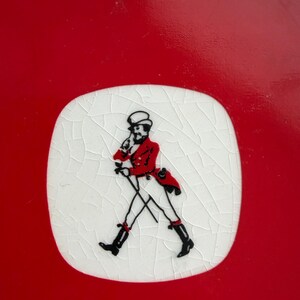 May include: A red and white ceramic coaster featuring a black and red illustration of a man walking with a cane. The illustration is on a white, cracked surface, set against a solid red background. The coaster is likely for beverages.