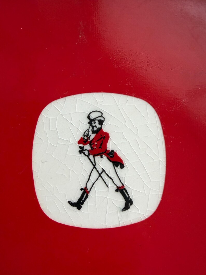 May include: A red and white ceramic coaster featuring a black and red illustration of a man walking with a cane. The illustration is on a white, cracked surface, set against a solid red background. The coaster is likely for beverages.