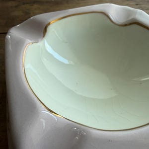 K&ouml;nnte beinhalten: Ein Vintage-Aschenbecher mit hellrosa Au&szlig;en- und cremefarbenem Innenbereich. Der Rand ist mit einem d&uuml;nnen Goldband akzentuiert. Der Aschenbecher hat eine rechteckige Form mit einer geschwungenen Kante.