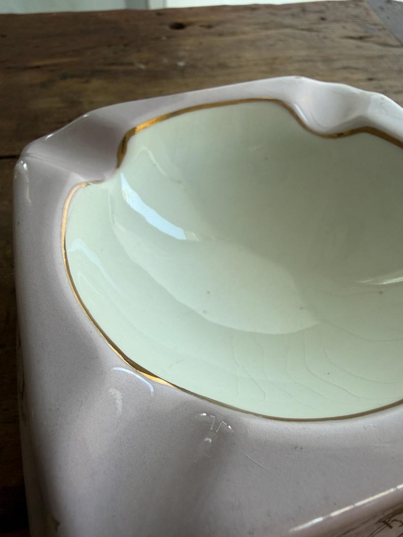 K&ouml;nnte beinhalten: Ein Vintage-Aschenbecher mit hellrosa Au&szlig;en- und cremefarbenem Innenbereich. Der Rand ist mit einem d&uuml;nnen Goldband akzentuiert. Der Aschenbecher hat eine rechteckige Form mit einer geschwungenen Kante.