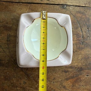 K&ouml;nnte beinhalten: Ein quadratischer, hellrosa Aschenbecher mit cremefarbenem Innenraum und goldenem Rand. Der Aschenbecher ist etwa 13 cm breit und 13 cm lang. Ein Ma&szlig;band liegt auf dem Aschenbecher.