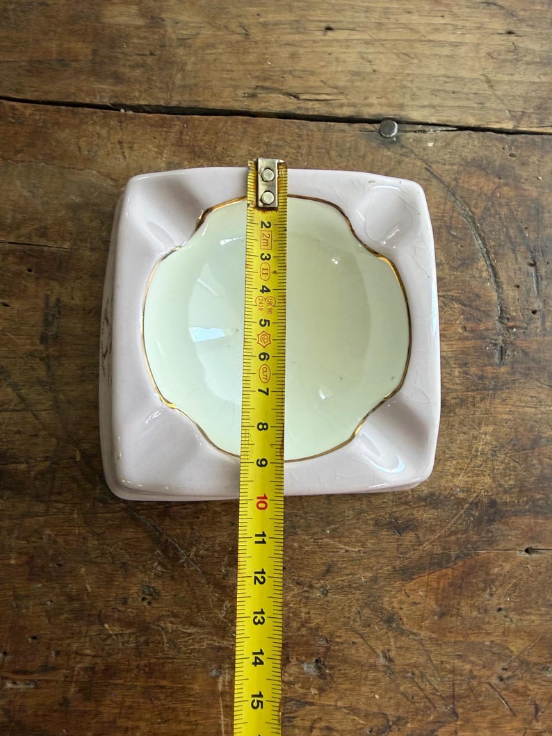 K&ouml;nnte beinhalten: Ein quadratischer, hellrosa Aschenbecher mit cremefarbenem Innenraum und goldenem Rand. Der Aschenbecher ist etwa 13 cm breit und 13 cm lang. Ein Ma&szlig;band liegt auf dem Aschenbecher.