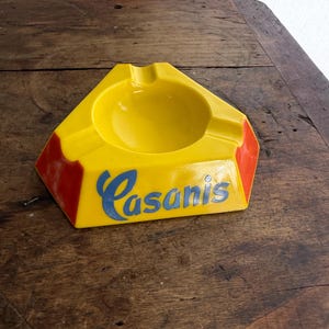 May include: Yellow ashtray with a triangular shape and a circular indentation. The ashtray has red accents on the corners and the word "Casanis" in blue script. A vintage smoking accessory.