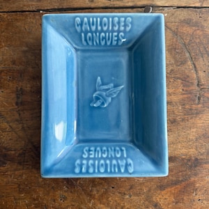 Op de afbeelding: Een rechthoekige, lichtblauwe keramische asbak met de woorden "CAULOISES LONGUES" in reliëf. In het midden bevindt zich een klein, in reliëf aangebracht ontwerp. De asbak staat op een houten oppervlak.