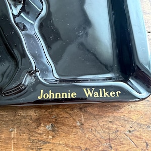 Pode incluir: Cinzeiro preto com o texto "Johnnie Walker" em letras douradas. O cinzeiro tem formato retangular com cantos arredondados e uma borda elevada. Possui acabamento brilhante.