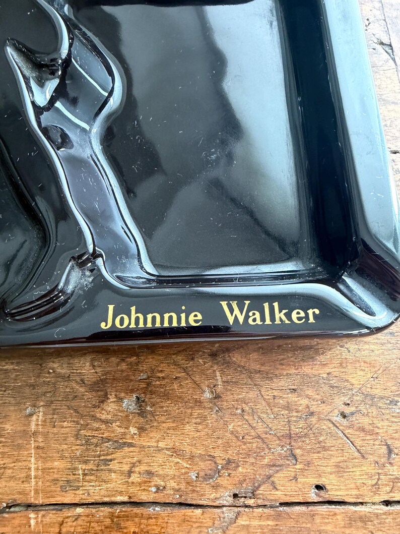 Pode incluir: Cinzeiro preto com o texto "Johnnie Walker" em letras douradas. O cinzeiro tem formato retangular com cantos arredondados e uma borda elevada. Possui acabamento brilhante.