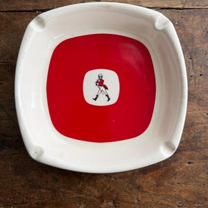 May include: Square ashtray with a white rim and a red center. The center features a small white square with a black and red illustration of a man walking. The ashtray has four small indentations for resting cigarettes.