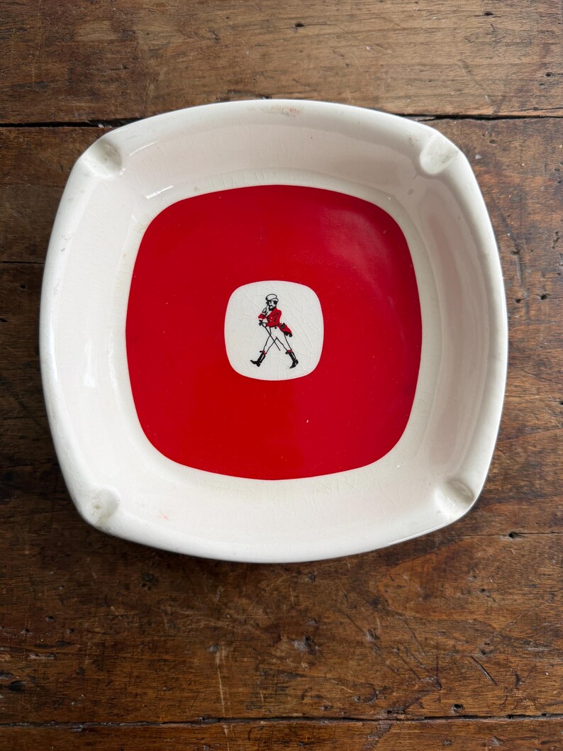 May include: Square ashtray with a white rim and a red center. The center features a small white square with a black and red illustration of a man walking. The ashtray has four small indentations for resting cigarettes.