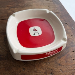 May include: Square, white ceramic ashtray with a red centre. The centre features a small white square with a figure in red. The ashtray has the words "Johnnie Walker" in red.