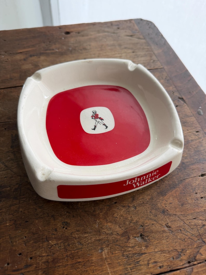 May include: Square, white ceramic ashtray with a red center. The center features a small white square with a figure in red. The ashtray has the words "Johnnie Walker" in red.