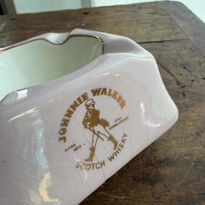 K&ouml;nnte beinhalten: Ein vintage, rosa Aschenbecher mit goldenem Rand und dem Logo "Johnnie Walker Scotch Whisky". Das Logo zeigt eine laufende Figur und Text. Der Aschenbecher hat eine rechteckige Form mit gebogenen Seiten.