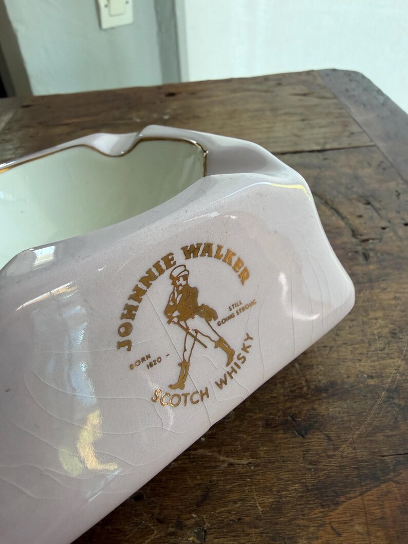 K&ouml;nnte beinhalten: Ein vintage, rosa Aschenbecher mit goldenem Rand und dem Logo "Johnnie Walker Scotch Whisky". Das Logo zeigt eine laufende Figur und Text. Der Aschenbecher hat eine rechteckige Form mit gebogenen Seiten.
