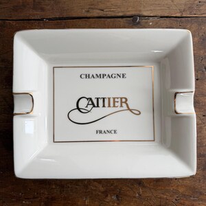 May include: White rectangular ashtray with gold trim and two cigarette rests. The center features the word "CHAMPAGNE" above the name "CATTIER" in elegant script, and "FRANCE" below. The ashtray is made of ceramic.