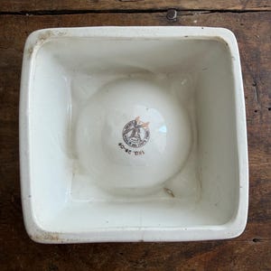 K&ouml;nnte beinhalten: Eine quadratische, cremefarbene Keramikschale mit abgerundeter Innenbasis. Die Schale hat einen leicht erh&ouml;hten Rand und ein verblasstes braun-wei&szlig;es Emblem in der Mitte. Der Text "TRU. 28-09" ist auf dem Emblem zu sehen.