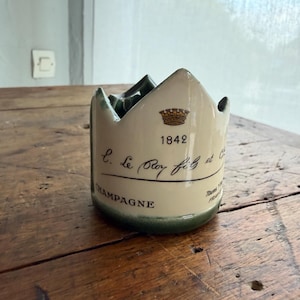 Peut inclure: Un porte-objet décoratif fabriqué à partir d'une bouteille de champagne. La céramique crème présente un emblème de couronne, l'année 1842 et le mot « CHAMPAGNE » en écriture élégante. Le support est posé sur une surface en bois.