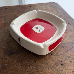 May include: A square ashtray with a red center and white sides. The center features a small graphic of a person in a red outfit. The sides have a red stripe and a small graphic of a person in a red outfit.