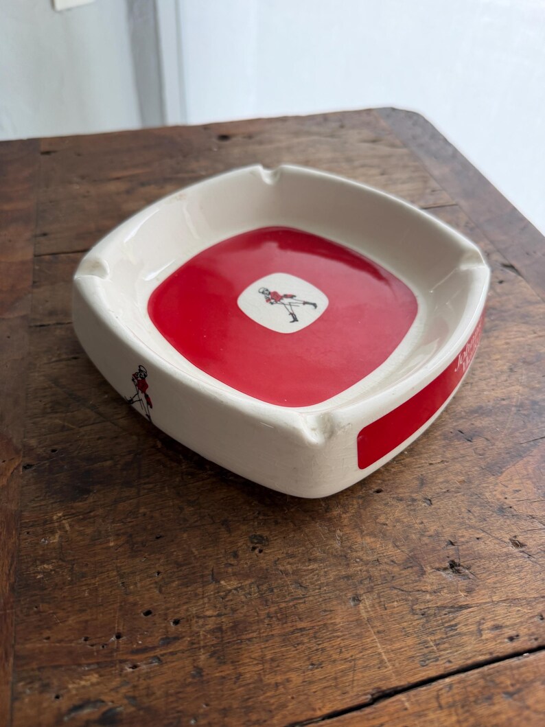 May include: A square ashtray with a red center and white sides. The center features a small graphic of a person in a red outfit. The sides have a red stripe and a small graphic of a person in a red outfit.